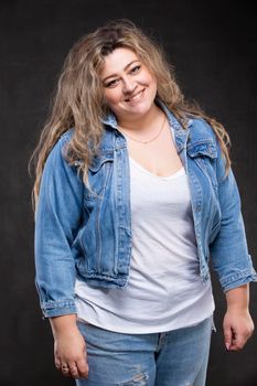 A beautiful fat woman in a denim jacket looks at the camera and smiles on a gray background.