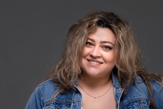 Portrait of a beautiful fat woman in a denim jacket looking at the camera and smiling on a gray background.