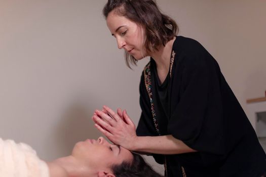 A masseuse massages the face of her patient who is lying face up on the table to relieve her health issues and muscle pain