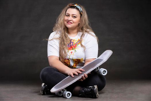 Beautiful fat woman with a skateboard on a gray background looks at the camera and smiles.