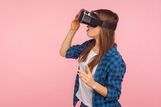 Side view of gamer girl in checkered shirt wearing vr headset, playing virtual reality game with amazed facial expression, surprised by innovative technology. studio shot isolated on pink background