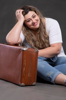 Beautiful fat woman in denim clothes with an old retro suitcase on a gray background. Plus size model girl.