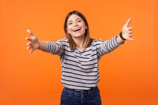 Come into my arms. Portrait of friendly positive woman with brown hair in casual shirt standing with stretched arms smiling at camera, going to hug. indoor studio shot isolated on orange background