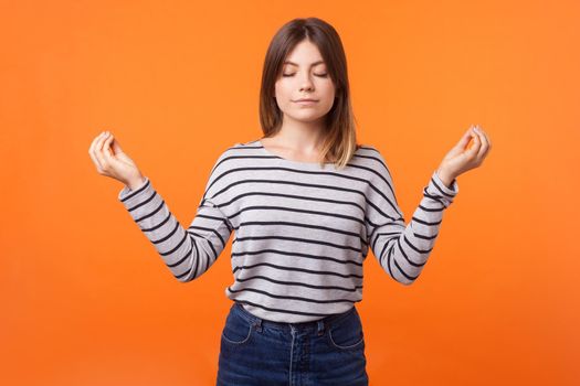 Portrait of calm young woman with brown hair in long sleeve striped shirt standing with closed eyes, meditating with mudra gesture, peaceful mind. indoor studio shot isolated on orange background