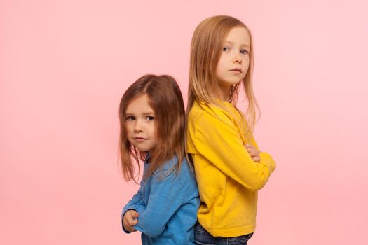 Friendship, togetherness and support. Two wonderful preschool girls standing back to back, looking at camera with confident attentive serious expression. indoor studio shot isolated on pink background