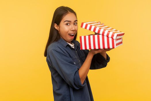 Portrait of happy amazed girl in denim shirt opening gift and looking at camera with surprised expression, excited joyful emotions, shocked by present. indoor studio shot isolated on yellow background