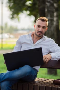 A business man in a white shirt is working in the park with a laptop. A young man on a background of green trees, a hot sunny summer day. Warm soft light, close-up.