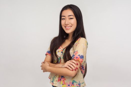 Portrait of calm friendly kind asian girl in summer blouse standing with crossed hands and looking at camera with toothy smile, optimistic outlook. indoor studio shot isolated on gray background