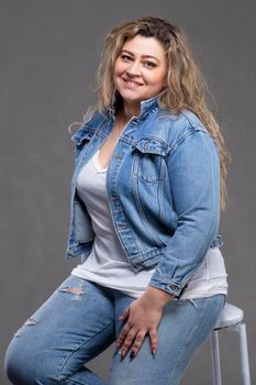 A beautiful fat woman in a denim jacket looks at the camera and smiles on a gray background.