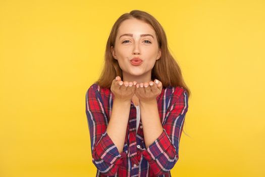 Portrait of beautiful lovely ginger girl in red shirt sending sensual air kiss with pout lips and flirting, looking with affectionate eyes, love feelings. studio shot isolated on yellow background