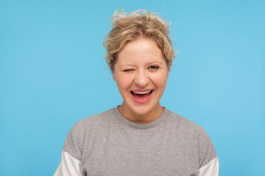 Carefree playful woman with short curly hair in grey sweatshirt winking at camera and smiling joyfully, flirting, expressing optimism and happiness. indoor studio shot isolated on blue background