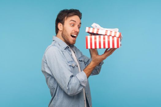 Portrait of amazed man in denim shirt opening gift, peeking inside box with surprised look, unpacking present, shocked by unbelievable birthday surprise. indoor studio shot isolated on blue background