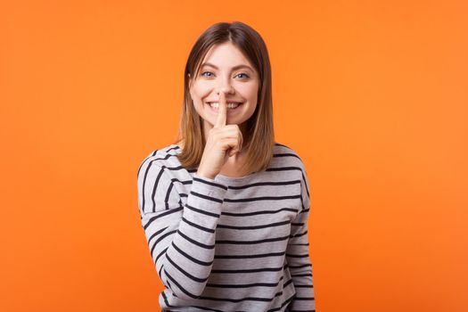 Portrait of positive beautiful woman with brown hair in long sleeve striped shirt standing, showing hush sign and looking at camera, sharing secret. indoor studio shot isolated on orange background