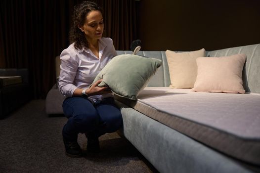 African American sales assistant, consultant in a designer furniture store, straightens a green velour pillow and dusts off a comfortable modern new upholstered sofa bed with orthopedic mattress