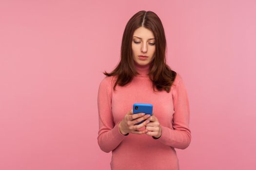 Serious brunette woman holding smartphone in hands, chatting with friends or making food order, online services. Indoor studio shot isolated on pink background