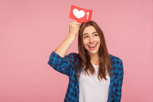 Portrait of beautiful positive blogger girl in checkered shirt dreaming of more likes in social media, holding heart icon on head, thinking over trendy interesting content. studio shot pink background