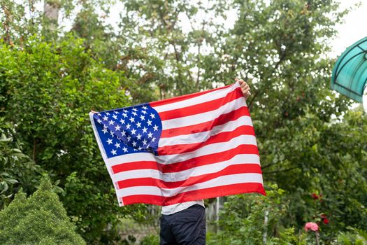 Positive attractive american young guy wrapped himself in the flag of America and looks at the camera, outdoors. High quality photo