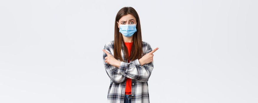 Coronavirus outbreak, leisure on quarantine, social distancing and emotions concept. Indecisive and clueless pretty girl in medical mask ask your opinion, advice as pointing sideways left and right.