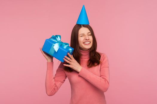 Happy funny brunette woman in party cap holding and shaking blue gift box trying to guess what is inside, birthday surprise. Indoor studio shot isolated on pink background