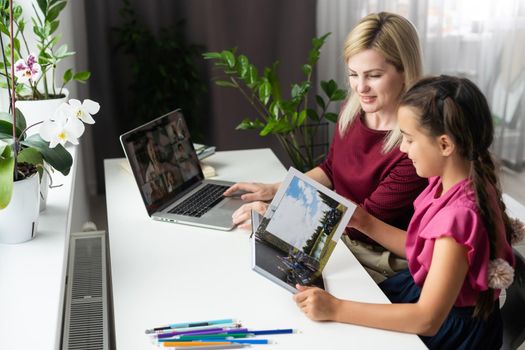 Pleased mother and small daughter sitting at table and school supplies and laptop and doing home task together in apartment.