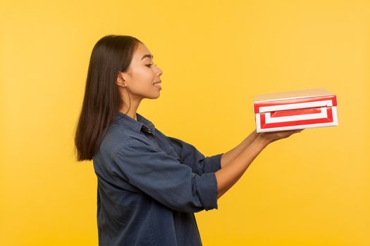 Take this present. Side view of generous happy brunette girl in denim shirt giving box and smiling, sharing holiday gift, congratulating on birthday. indoor studio shot isolated on yellow background