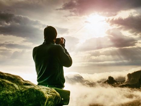 Photographer on cliff with camera viewfinder in his face. Nature photographer takes photos with mirror camera on peak of rock. Dreamy fogy landscape 