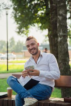 A happy man is sitting on a bench in the park, counting dollars. A young man on a background of green trees, a hot sunny summer day. Warm soft light, close-up.