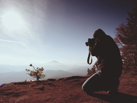 Photographer down on knees takes photos with mirror camera on peak of rock. Dreamy landscape, orange Sun at horizon