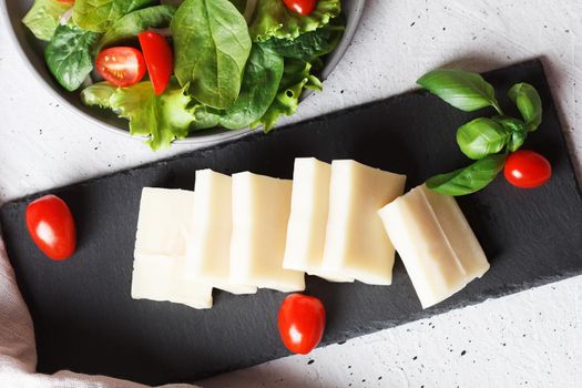 A piece of fresh sliced halloumi on a slate board with cherry tomatoes and spinach leaves. Preparing to cook cheese on the grill. Top view