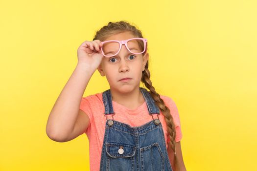 Portrait of amazed stylish little girl in denim overalls taking off pink eyeglasses and looking with astonished expression, child surprised by her vision improvement. indoor studio shot, isolated