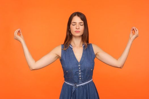 Zen practice, breath technique. Brunette woman in denim dress doing yoga exercise, holding hands in mudra gesture and meditating, feeling tranquil relaxed. studio shot isolated on orange background