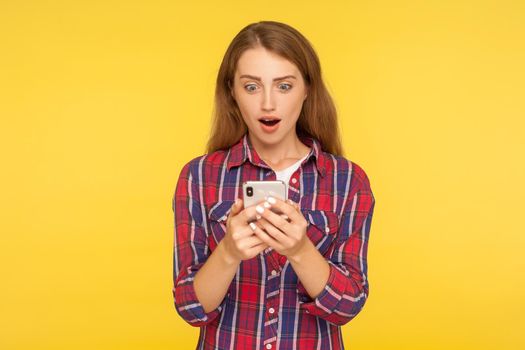 Convenient online app. Portrait of amazed ginger girl using mobile phone and looking shocked pleasantly surprised by mobile service, typing message. indoor studio shot isolated on yellow background