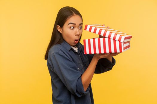 Portrait of amazed funny girl in denim shirt opening gift and looking into box with shocked astonished expression, found unbelievable present surprise. indoor studio shot isolated on yellow background