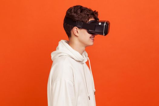 Profile portrait of happy young man in casual style hoodie enjoying view in virtual reality glasses, modern technologies. Indoor studio shot isolated on orange background