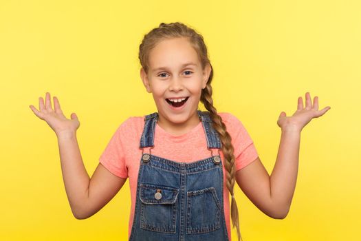Unexpected surprise. Portrait of astonished child in denim overalls standing with raised hands in amazement, shocked by sudden unbelievable news. indoor studio shot isolated on yellow background
