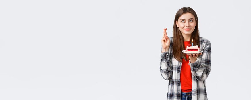 People lifestyle, holidays and celebration, emotions concept. Dreamy hopeful birthday girl in casual clothes, cross fingers and look up making wish, holding b-day cake, blowing candle.