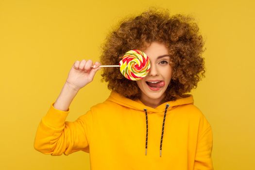 Portrait of funny modern curly-haired woman in urban style hoodie covering eye with lollipop and showing tongue out, playing with sweet rainbow candy. indoor studio shot isolated on yellow background