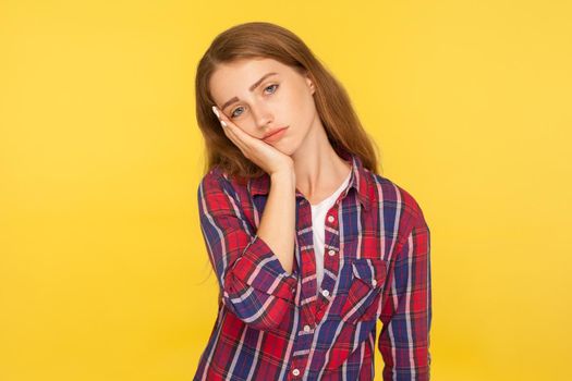 I'm bored. Portrait of depressed ginger girl in checkered shirt leaning on cheek and looking with indifferent dull expression, tired of boring story. indoor studio shot isolated on yellow background