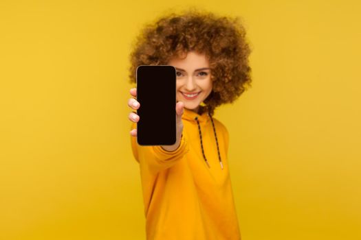 Portrait of cheerful curly-haired woman with engaging smile showing cellphone with empty display, mock up for app advertise, mobile web service. indoor studio shot isolated on yellow background