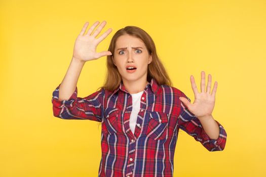 I'm afraid. Portrait of scared panicked ginger girl in checkered shirt making frightened gesture with raised hands, looking terrified and shocked. indoor studio shot isolated on yellow background