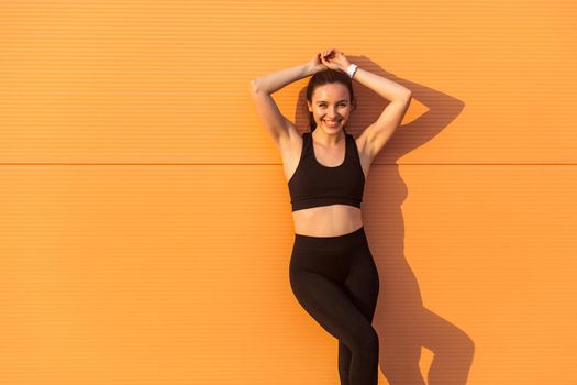 Beautiful sexy girl with fit body in tight sportswear, black pants and top, leaning against orange wall and smiling to camera, living healthy sport life, full of energy. isolated on advertising area