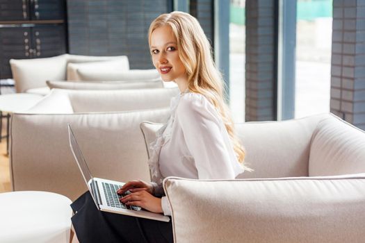 Portrait of stunning beautiful wealthy woman with blond hair in classic clothes sitting on comfortable couch holding laptop and smiling at camera, fashionable lady ready for online shopping. indoors