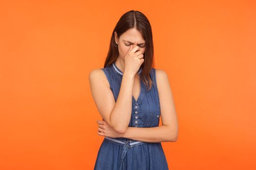 Frustrated sad brunette woman in denim dress crying, wiping away tears, feeling depressed helpless, upset about loss, bereavement, desperate emotions. indoor studio shot isolated on orange background