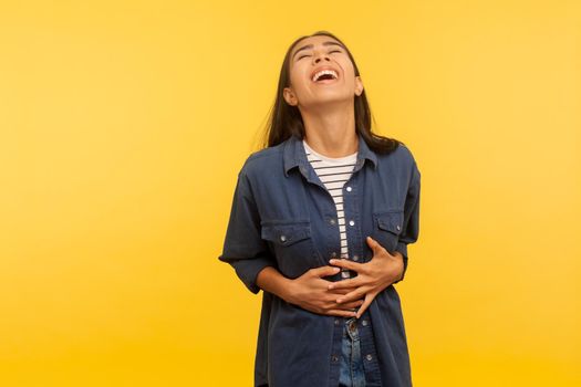 What a funny joke. Portrait of amused carefree girl in denim shirt cracking up with hilarious laughter, holding her stomach and laughing out loud. indoor studio shot isolated on yellow background