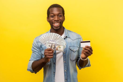 Portrait of excited young man in denim shirt holding dollar bills and credit card with satisfied expression, toothy smile, enjoying payments with cashback. studio shot isolated on yellow background