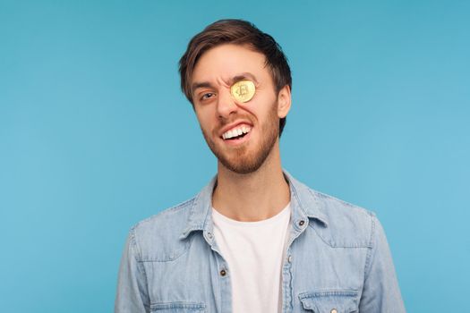 Cryptocurrency. Cheerful happy businessman in denim shirt standing with bitcoin placed in eye, smiling carefree and looking through golden btc coin. indoor studio shot isolated on blue background
