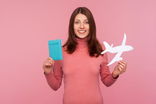Happy brunette woman in pink sweater holding paper plane model and passport in hands looking at camera with toothy smile. Indoor studio shot isolated on pink background