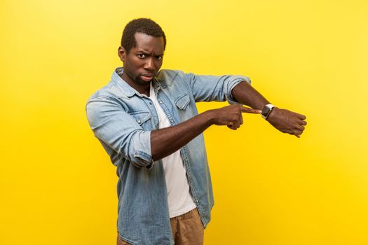 Look at the time. Portrait of irritated impatient man in denim casual shirt pointing at wristwatches, nervous about being late for meeting, deadline. indoor studio shot isolated on yellow background