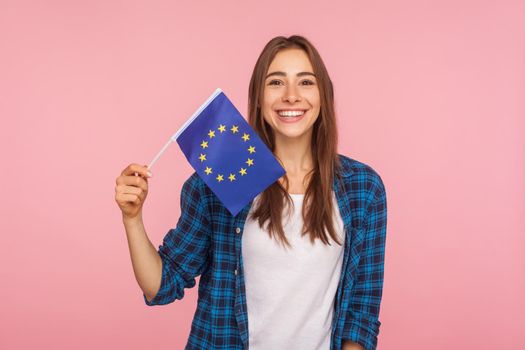 Portrait of friendly pretty woman in checkered shirt smiling broadly and holding flag of European Union, symbol of Europe, EU association and community. indoor studio shot isolated on pink background