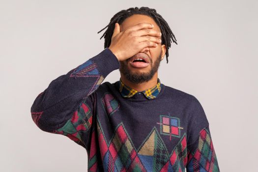 Angry shocked african guy with beard and dreadlocks closing eyes with hand, hiding, dont want to see and know, ignorance. Indoor studio shot isolated on gray background